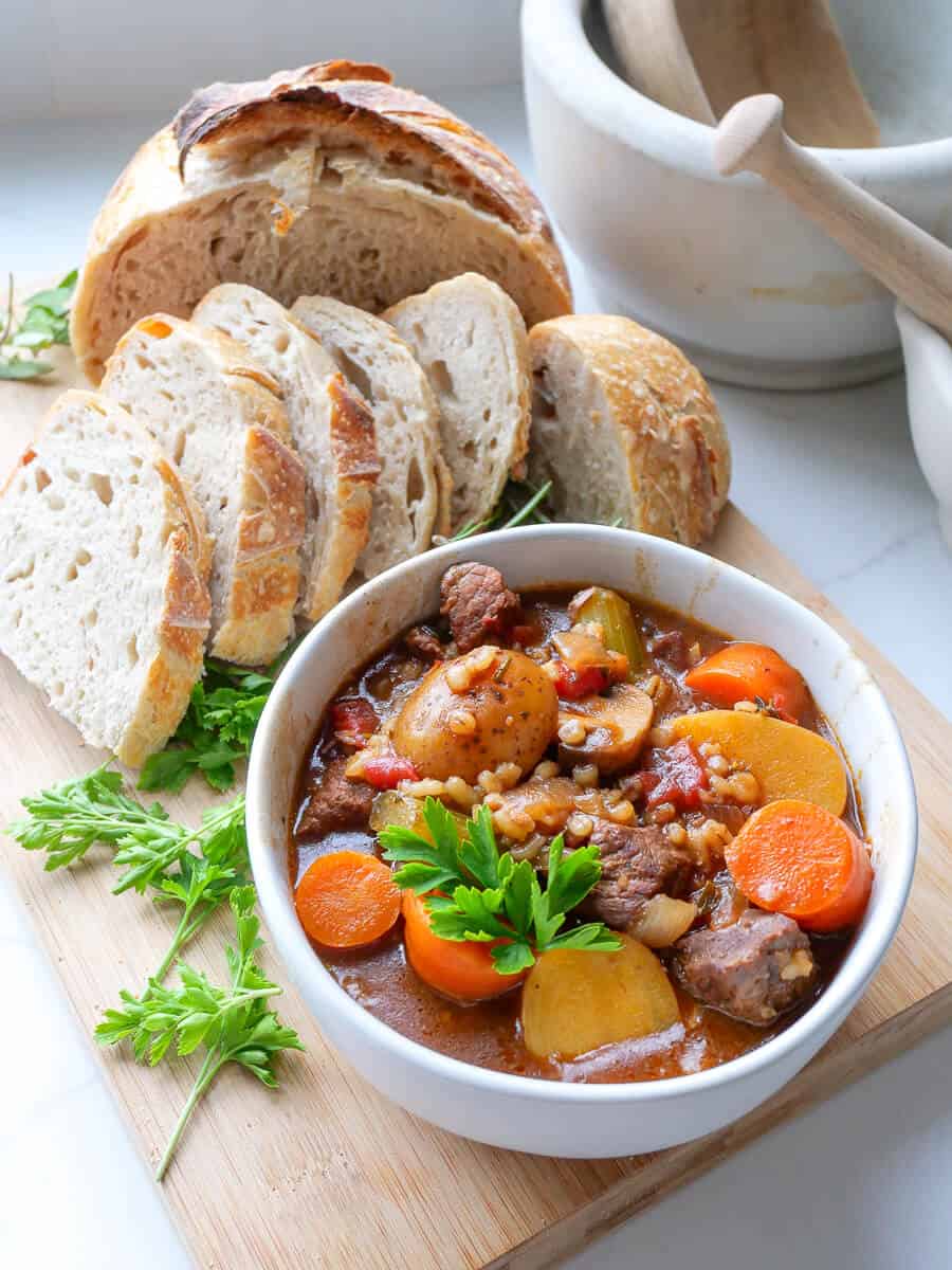 Bowl of beef barley stew on a counter with bread.