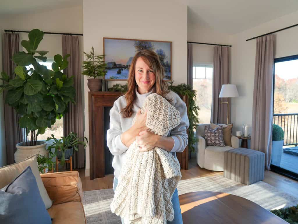 Woman holding a knit blanket in front of a cozy winter fireplace mantel.